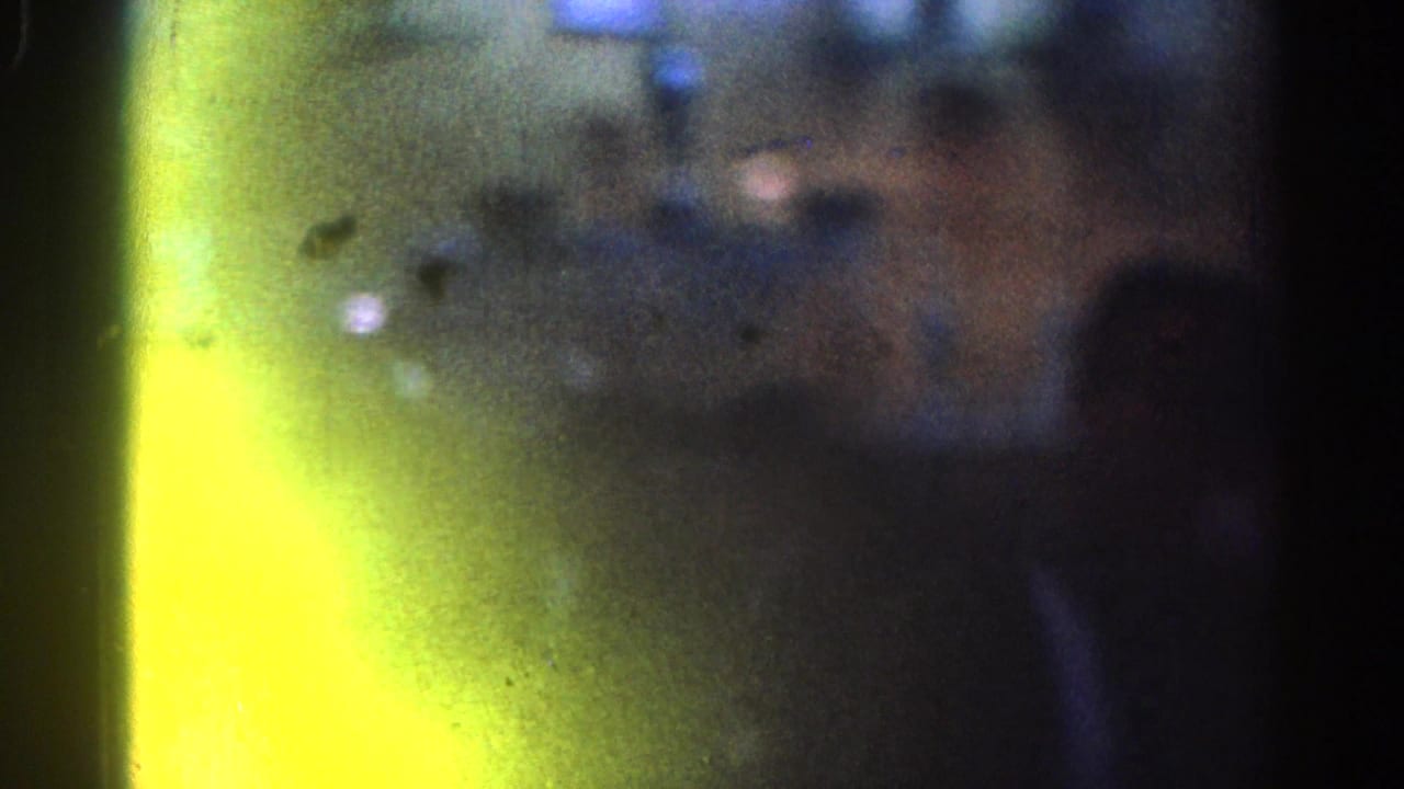 1967 Los Angeles 8mm frame with a yellow-green light leak, heavy grain, and shadowed indoor figures barely visible.