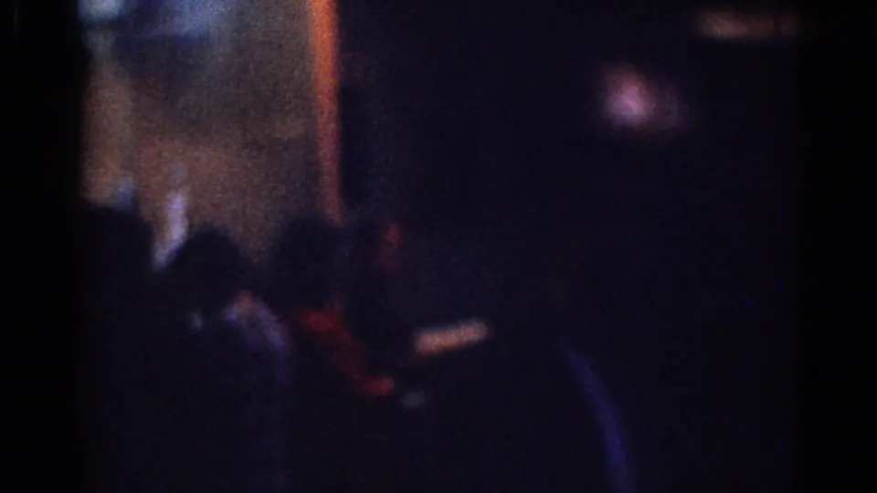 1967 Los Angeles 8mm frame showing an indistinct dark-suited person amid deep shadows and soft interior lights.