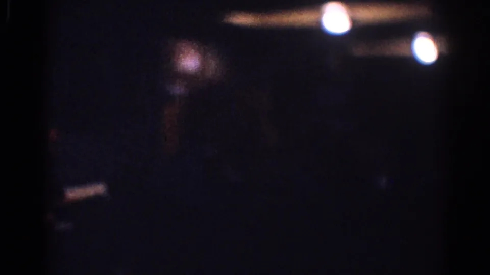 Low-light 1967 Los Angeles home-movie frame with soft ceiling lights, grain, and a barely visible person in shadow.