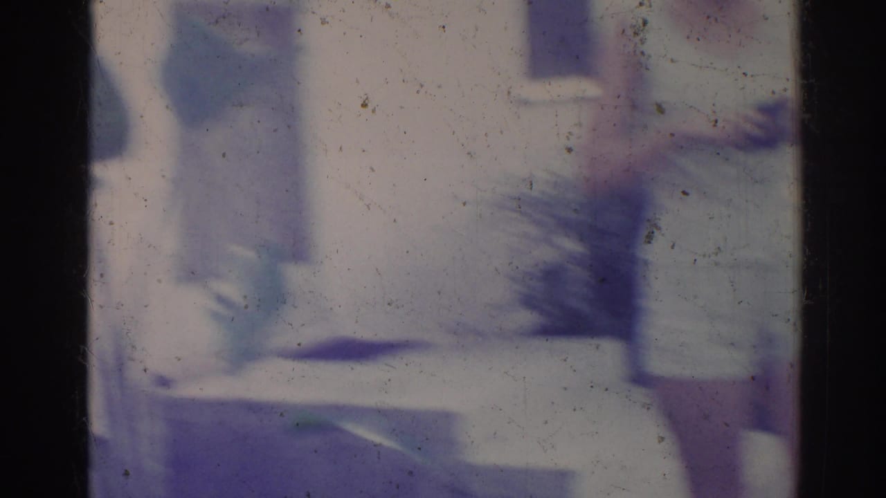 Aged 8mm frame showing a pale house exterior and indistinct leafy plants in soft focus, consistent with the Los Angeles home-movie setting.