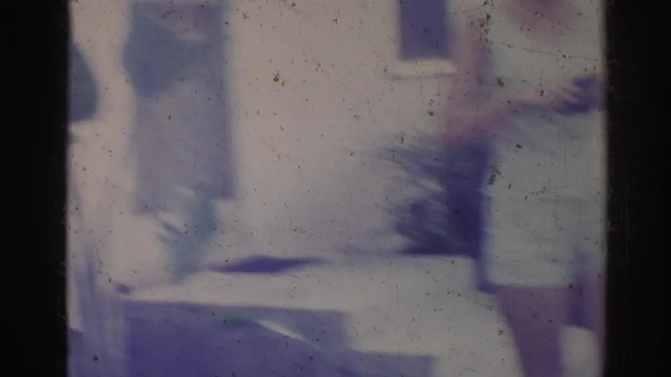 Aged 8mm frame showing a pale house exterior and indistinct leafy plants in soft focus, consistent with the Los Angeles home-movie setting.
