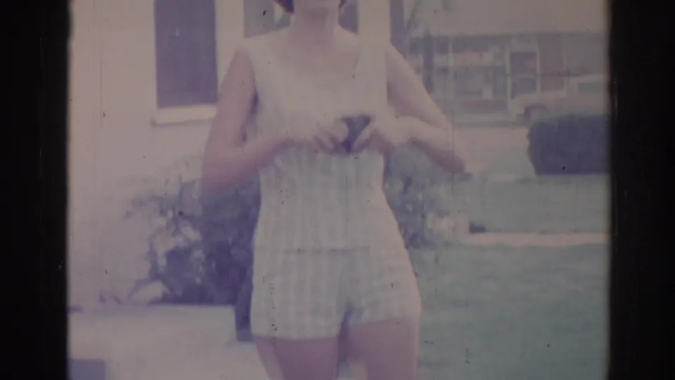 Woman in a light sleeveless dress or romper stands on a grassy Los Angeles lawn near a house and garden planting in this 1967 home movie frame.