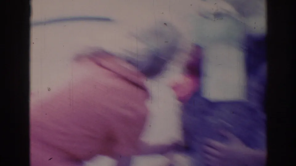 Motion-blurred 1967 8mm frame with a close, indistinct view of a person or clothing near plants and a pale house background.