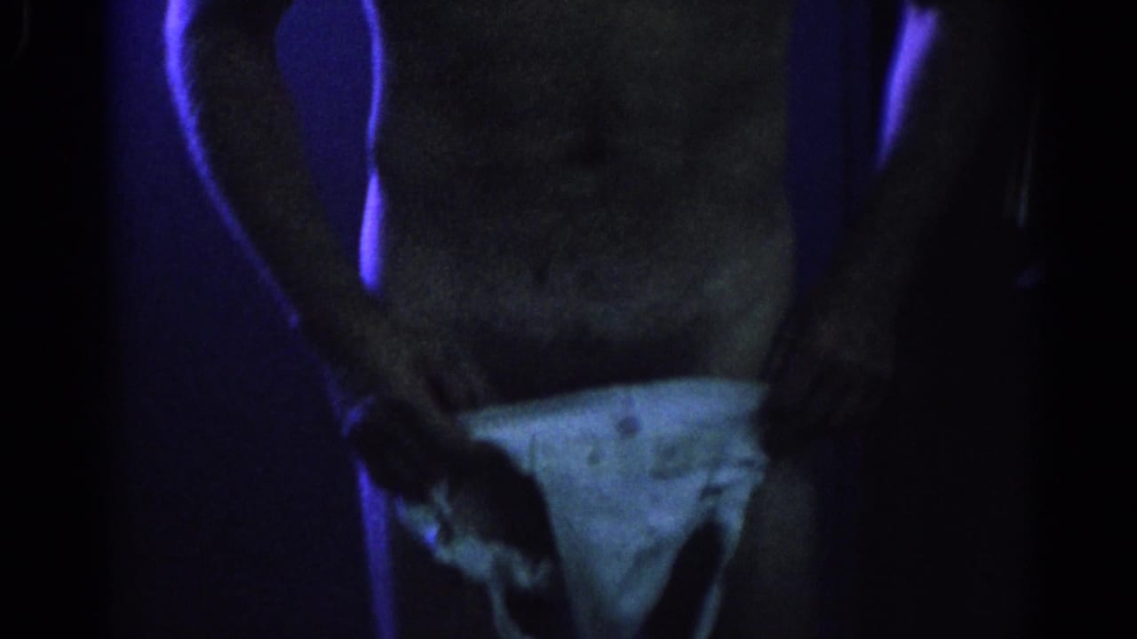 Man seen from torso to thighs wearing a white jockstrap in a dark room.