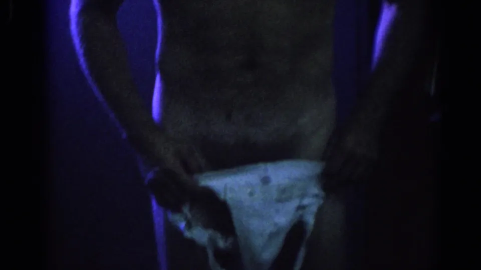 1967 Los Angeles 8mm frame of a man in a white jockstrap standing in a dim, blue-lit locker-room setting.