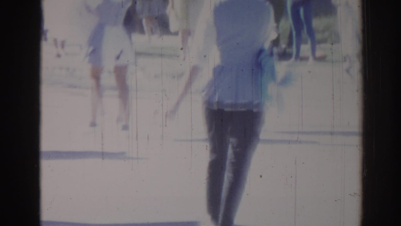 Blurred pedestrian walking in bright street scene with film scratches and dark frame edges.