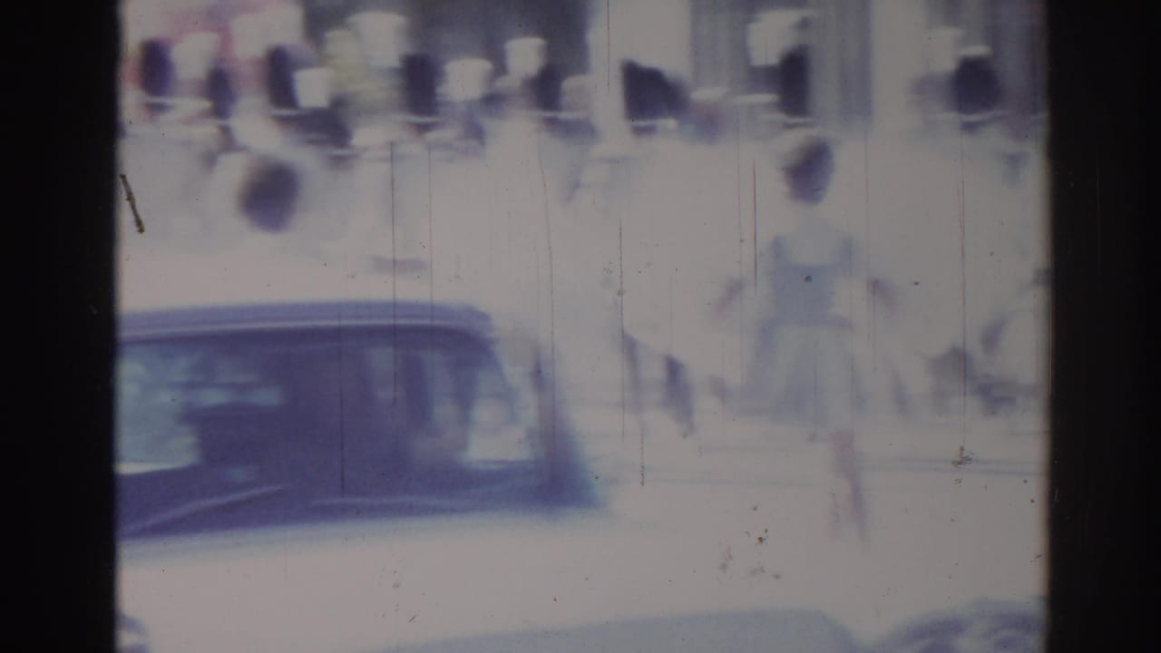1967 Los Angeles 8mm frame with a classic car passing below indistinct spectators and pedestrians at a street parade.