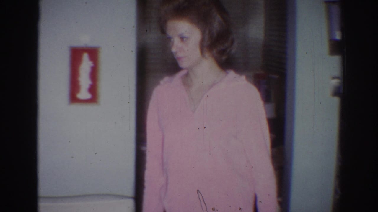 Woman in a pink hooded sweatshirt standing in a home interior near wall art.