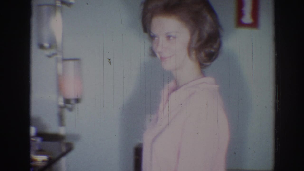 The woman smiles beside a small wall lamp in a private Los Angeles interior from 1967 8mm footage.