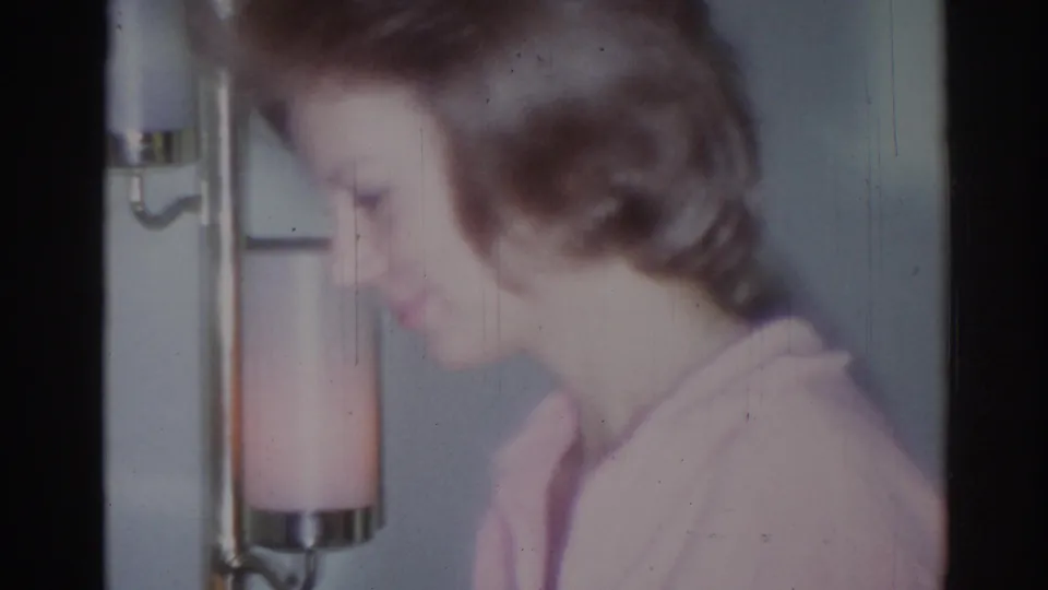 Soft-focus side view of the woman moving past a pink-toned lampshade in the 1967 Los Angeles home interior.