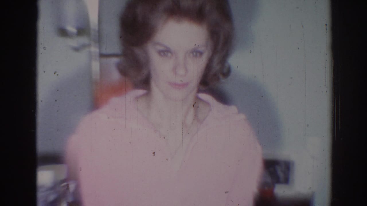 In a 1967 Los Angeles home-movie frame, an adult woman in a pink sweater faces the camera near a wall lamp and household furnishings.