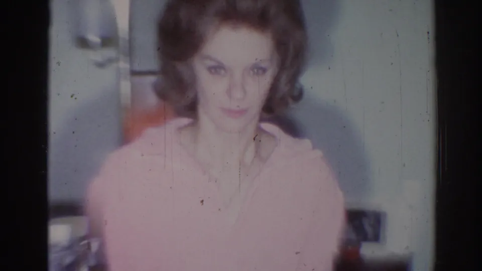 In a 1967 Los Angeles home-movie frame, an adult woman in a pink sweater faces the camera near a wall lamp and household furnishings.