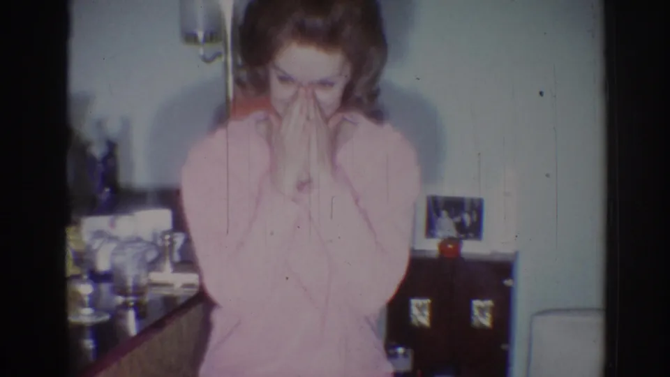 The woman in the pink sweater covers her mouth in a private Los Angeles interior, with glassware and framed photos visible behind her.
