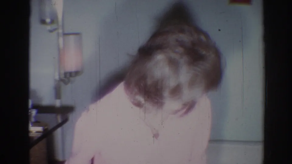 The woman bends forward so her face is partly obscured, with the pink sweater, tabletop, wall lamp, and pale interior wall still visible.