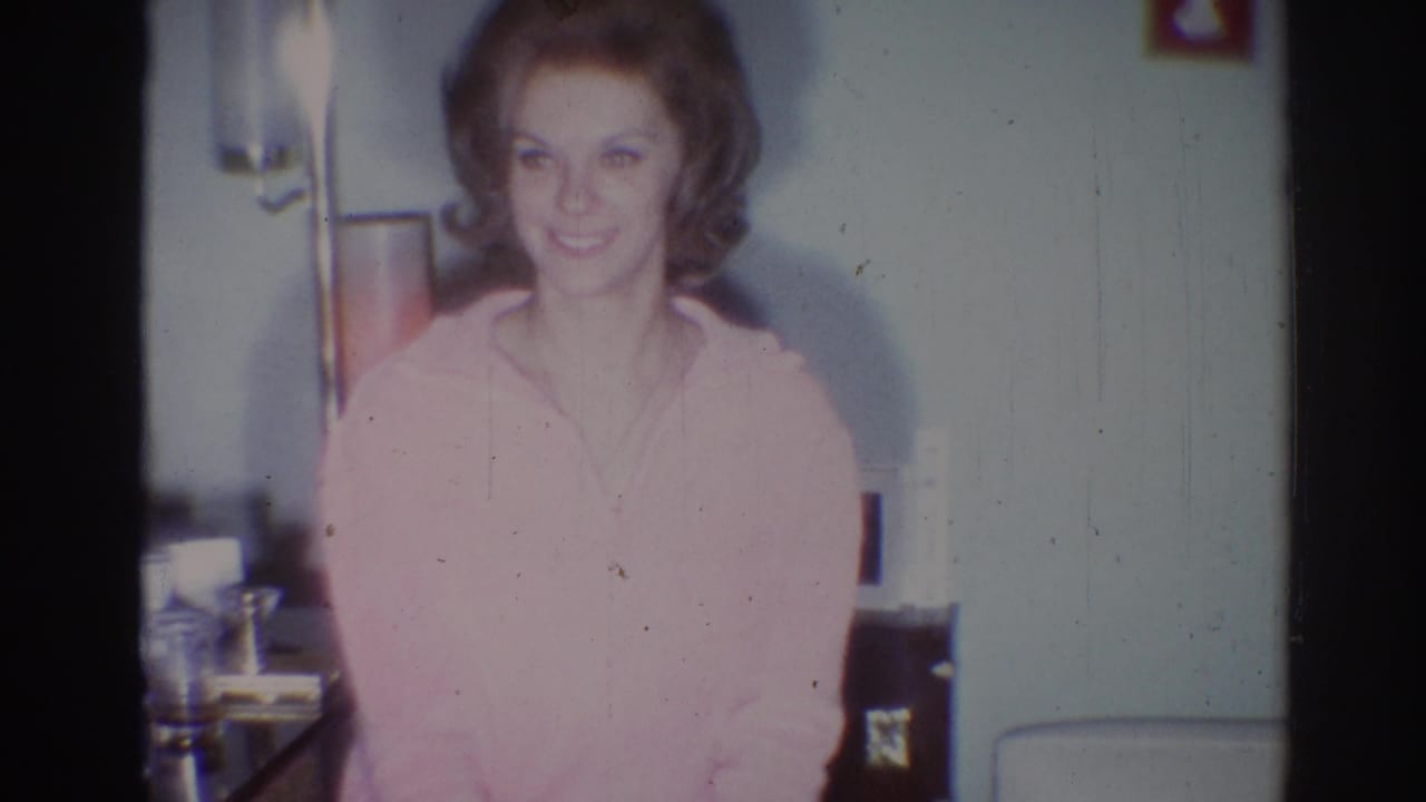 Young woman in a pink sweater smiles toward the camera in a domestic room.