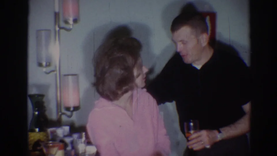 1967 Los Angeles home-movie frame of a man with a glass leaning toward a seated woman in a private room with candle sconces.