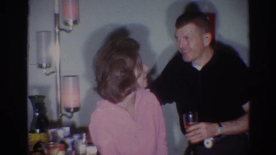 1967 Los Angeles 8mm frame of a smiling man holding a drink beside a woman in a dim private domestic interior.