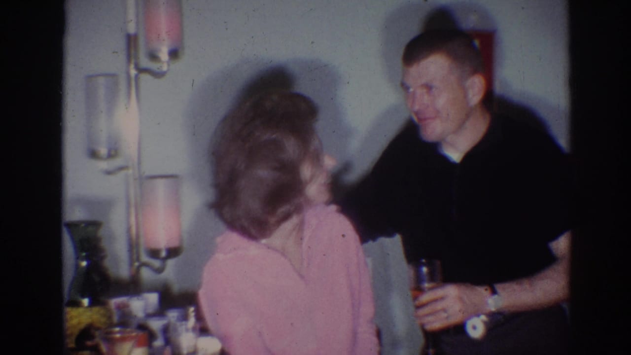 Woman in pink faces a smiling man in a dark shirt holding a drink beside a floor lamp.
