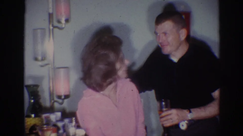 1967 Los Angeles home-movie frame of a woman in pink turning toward a smiling man holding a drink in a private interior.