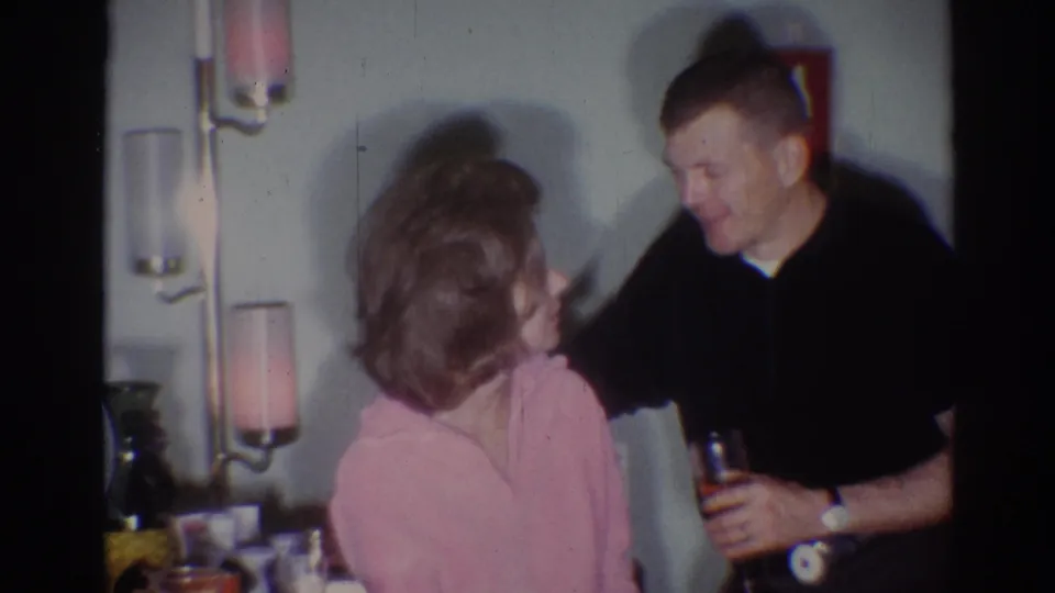 1967 Los Angeles home-movie frame with a woman in a pink top and bouffant hairstyle facing a man holding a drink indoors.
