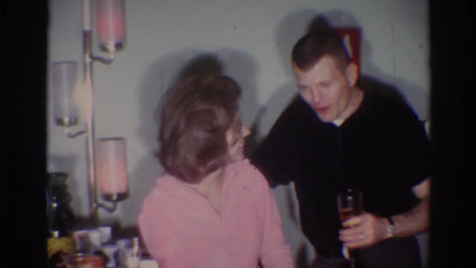 1967 Los Angeles home-movie frame of a man leaning toward a woman in pink while holding a drink during a casual indoor gathering.