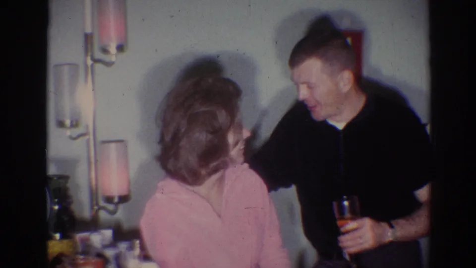 In 1967 Los Angeles home-movie footage, a man holding a drink leans toward a woman in a pink top inside a softly lit private room.