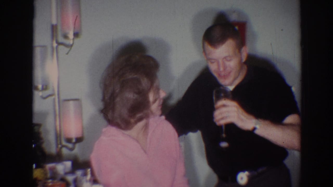 The man smiles with a drink in hand while the seated woman looks toward him in a 1967 Los Angeles private party interior.