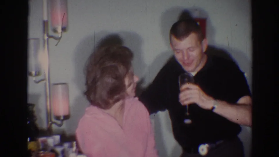 The man smiles with a drink in hand while the seated woman looks toward him in a 1967 Los Angeles private party interior.