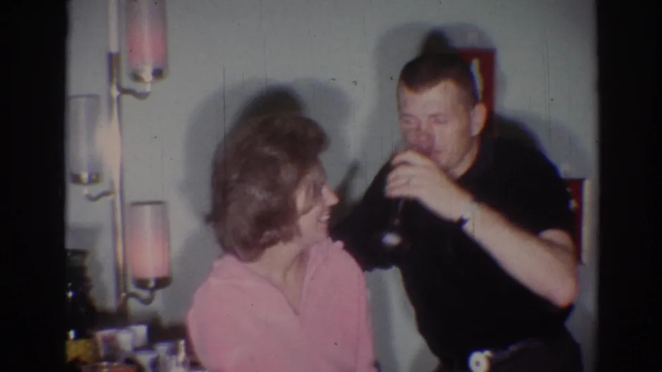 In the softly exposed 8mm frame, the man drinks from his glass while the woman in pink turns toward him at the party.