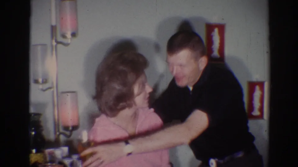 The man moves into a brief embrace or playful reach around the woman while holding a drink in the 1967 Los Angeles party footage.