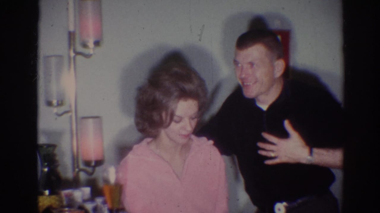 Woman in a pink robe sits beside a smiling man in a dark shirt indoors.