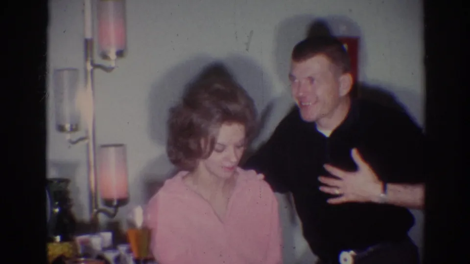 1967 Los Angeles home-movie frame showing a seated woman in a pink robe and a smiling man leaning close in a private interior.
