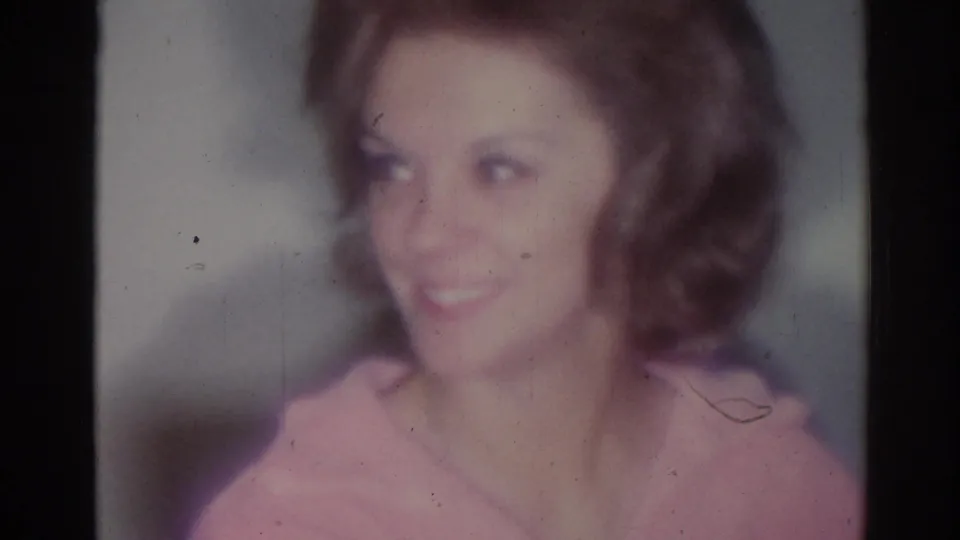Soft 1967 Los Angeles 8mm close-up of a woman in a pink top smiling inside a dim room.