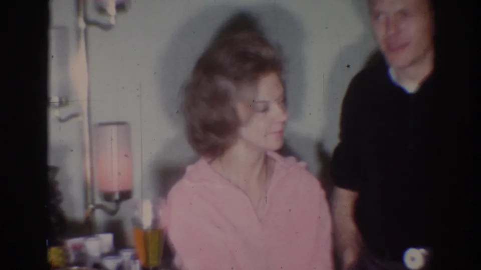 In a 1967 Los Angeles home interior, a seated woman in pink looks toward a standing man in a dark shirt.
