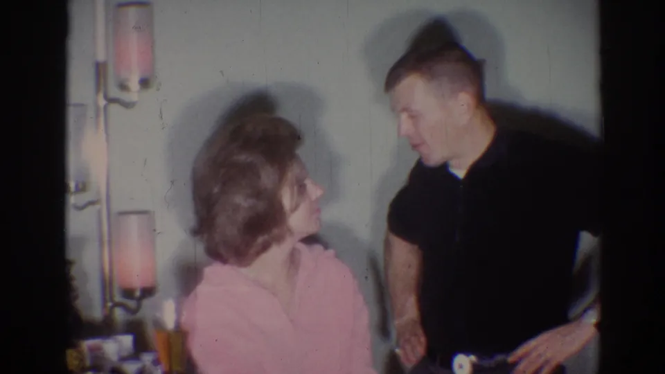 A man in a dark shirt leans toward a seated woman in pink during a 1967 Los Angeles home-movie conversation.