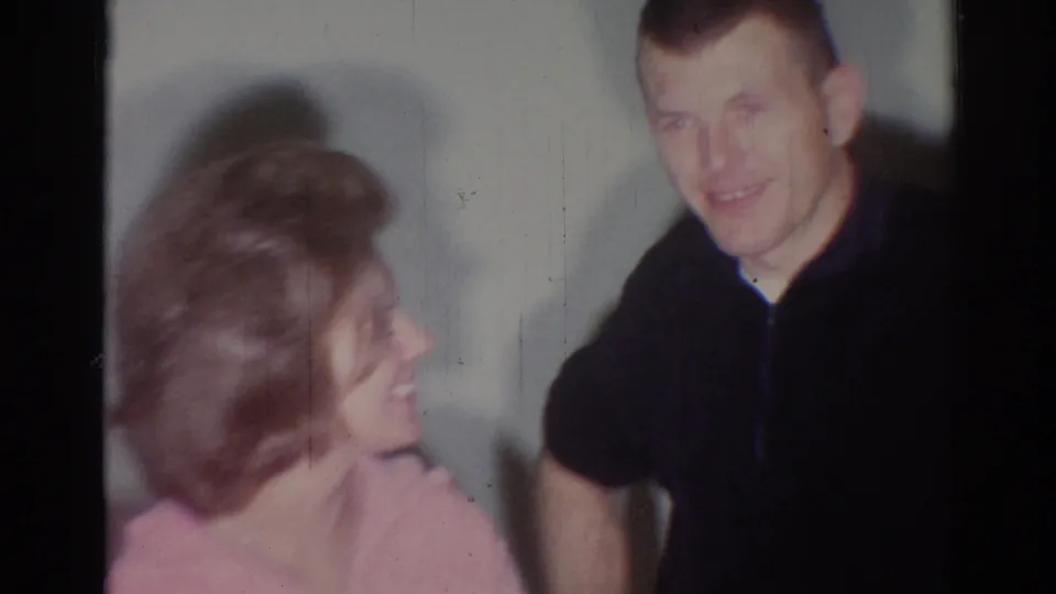 The woman smiles toward the man in a dark shirt in soft 1967 Los Angeles 8mm interior footage.