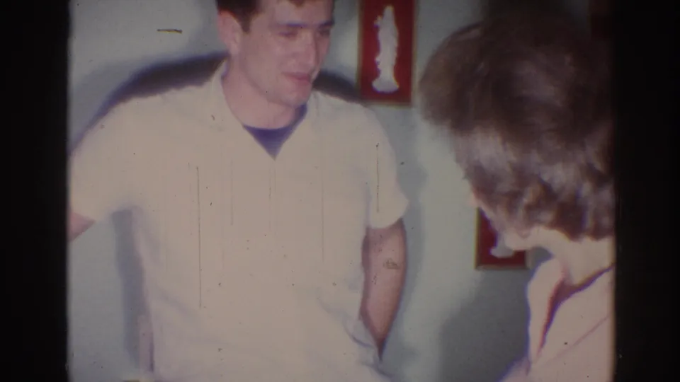 In a 1967 Los Angeles home-movie frame, a man in a pale short-sleeve shirt smiles while a woman with styled hair faces him indoors.