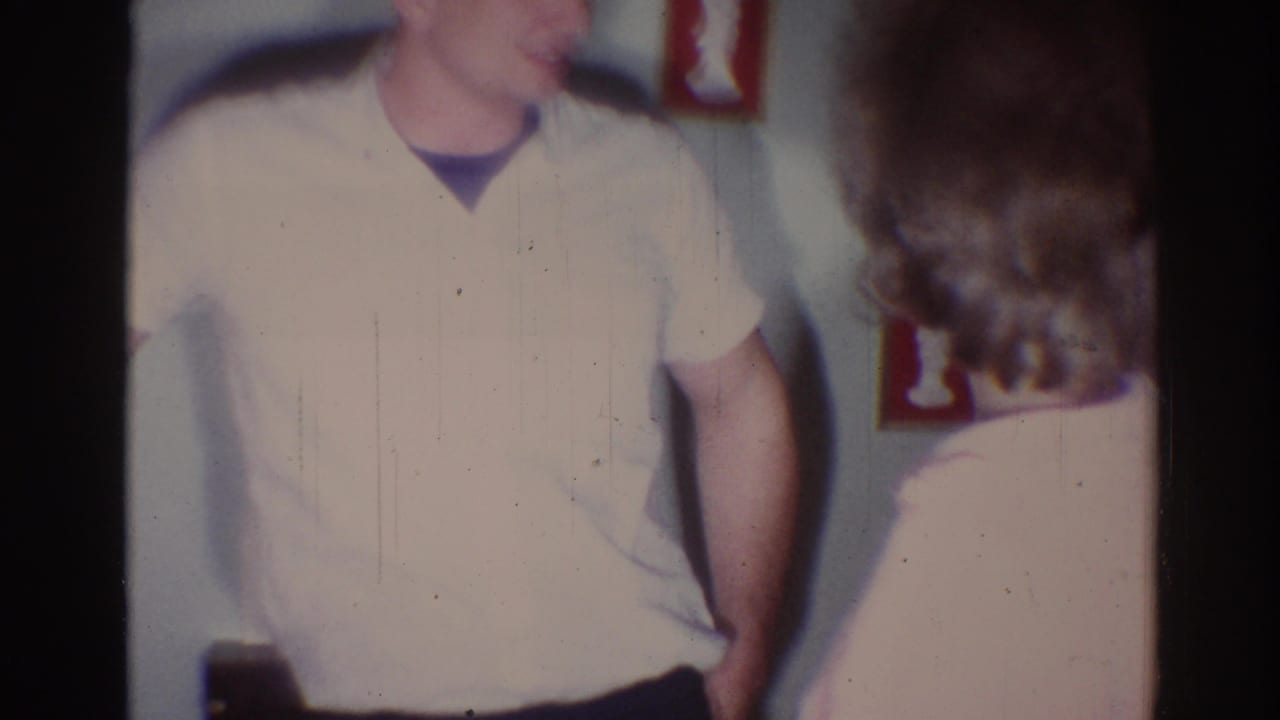 Man in a pale shirt stands near a woman in a dim home interior.