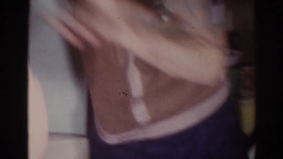 A blurred close-range 8mm frame shows a man in a brown vest gesturing with his hand inside the same domestic setting.