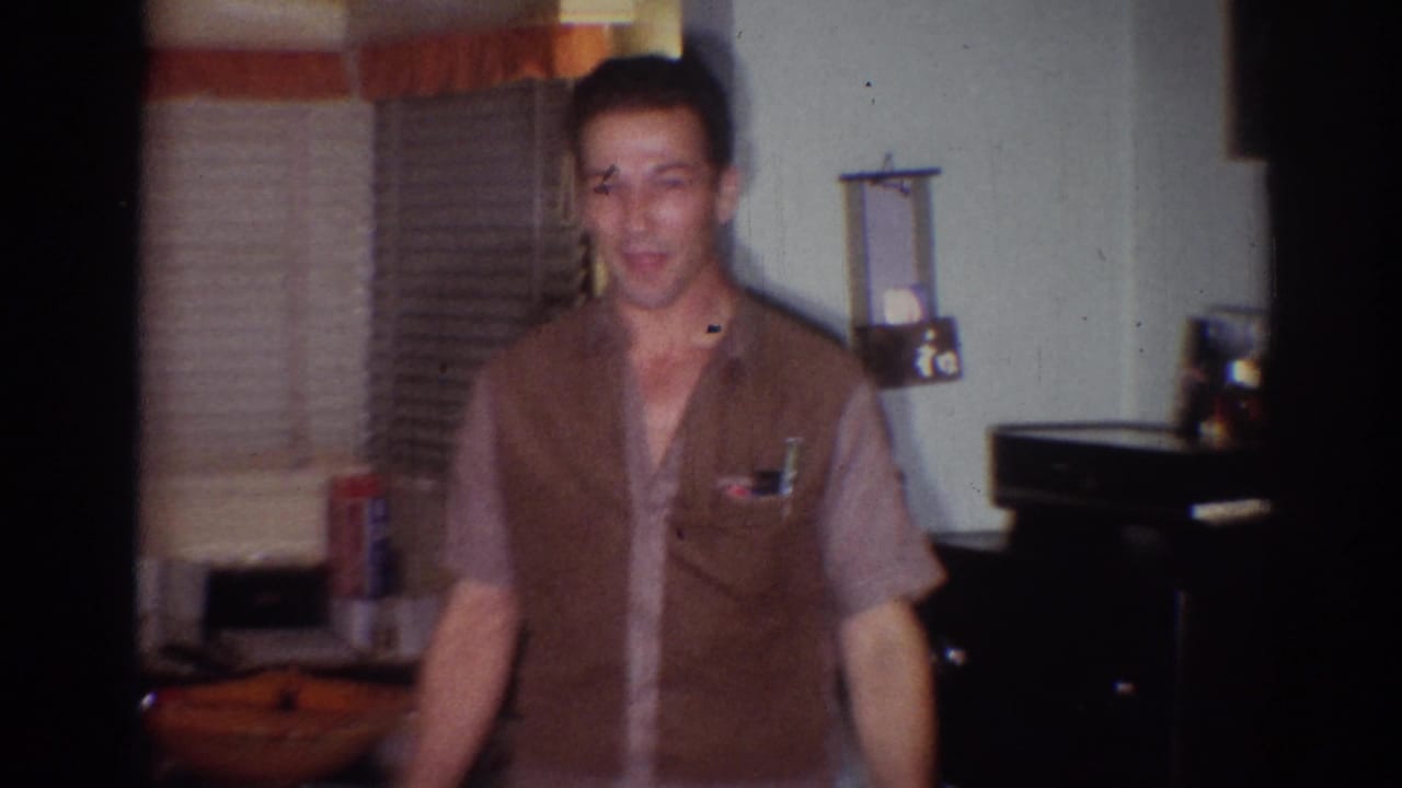 Man in a brown vest stands in a room with blinds and a lamp shade.