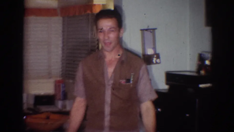 Adult man in a brown vest and light shirt stands in a dim Los Angeles room with blinds and household items visible.