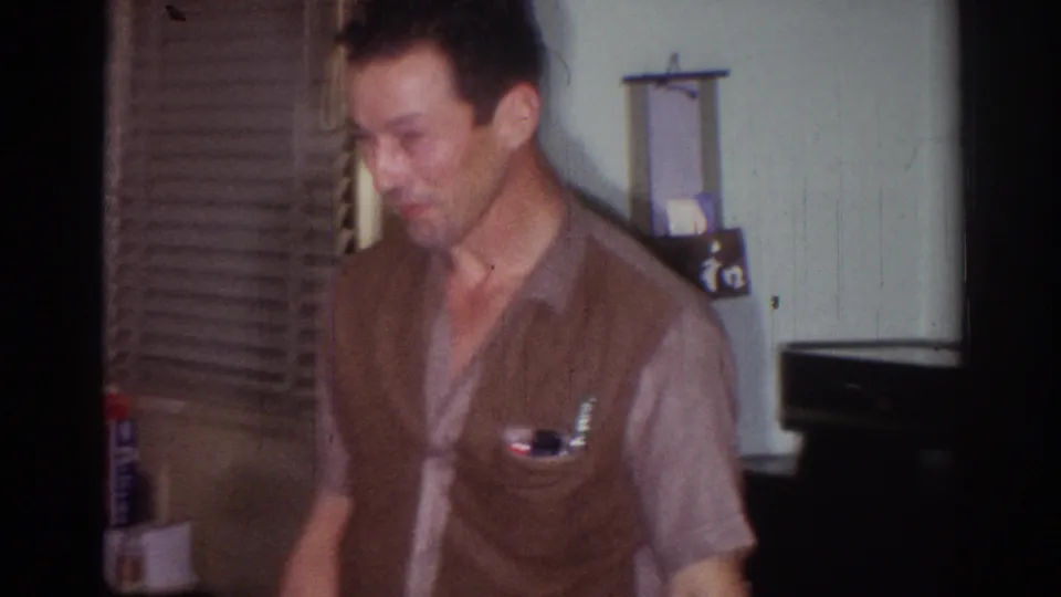 The man in a brown vest looks downward in a softly focused 1967 home-movie interior in Los Angeles.
