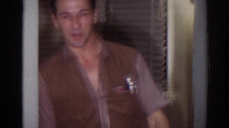 Close, softly focused frame of the man in a brown vest inside a dim 1967 Los Angeles home-movie interior.