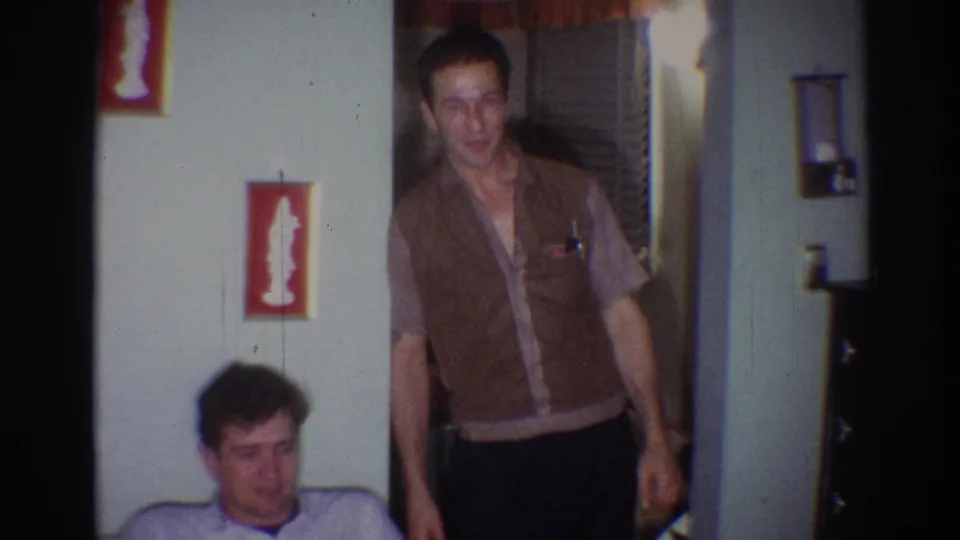 A standing man in a brown vest is seen near a seated man, with red wall decor and household furnishings visible in the room.