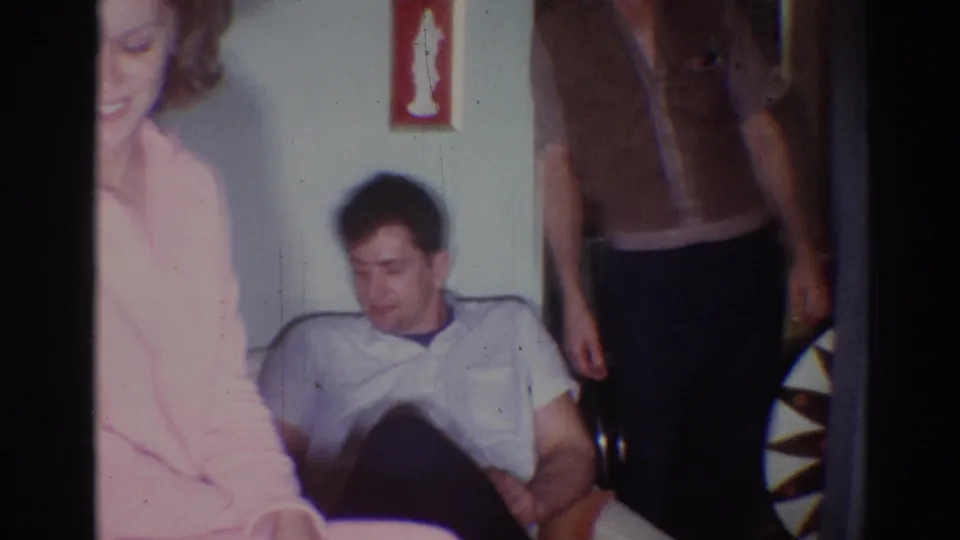 A seated man, a standing man in a brown vest, and a partially visible woman share the frame in a 1967 Los Angeles home interior.