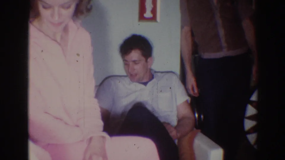 1967 Los Angeles home-movie frame showing a young man on a white couch with a woman in pink and another adult partly visible.