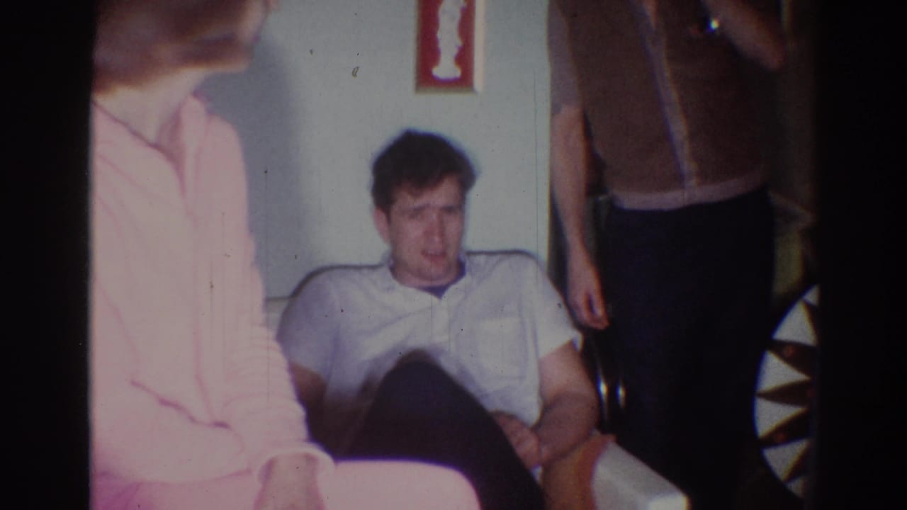 Seated man on a white couch with adults standing and sitting around him.