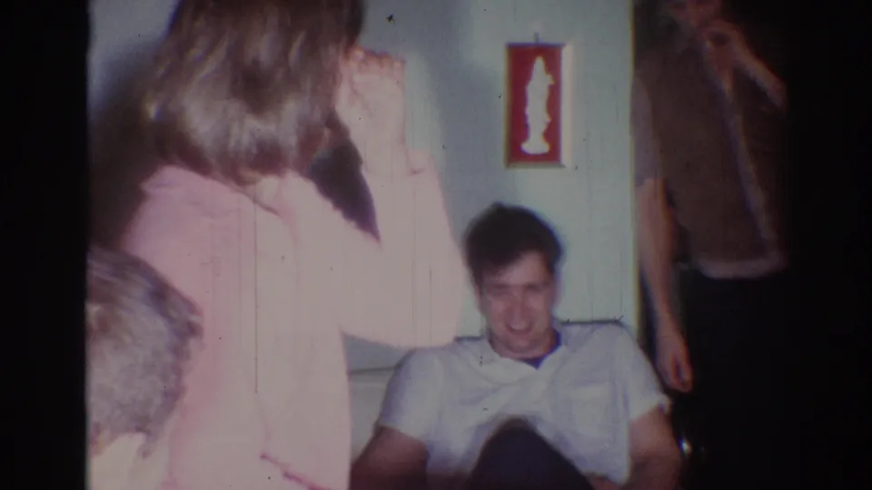 1967 Los Angeles home interior with a woman in pink in the foreground and a smiling man seated on the couch behind her.