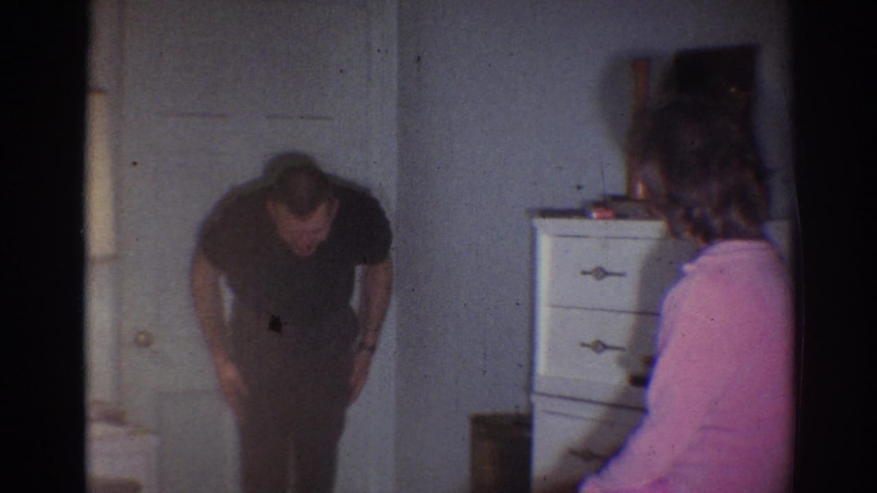 Man bends forward near a closed door while a woman in pink sits at the right edge.