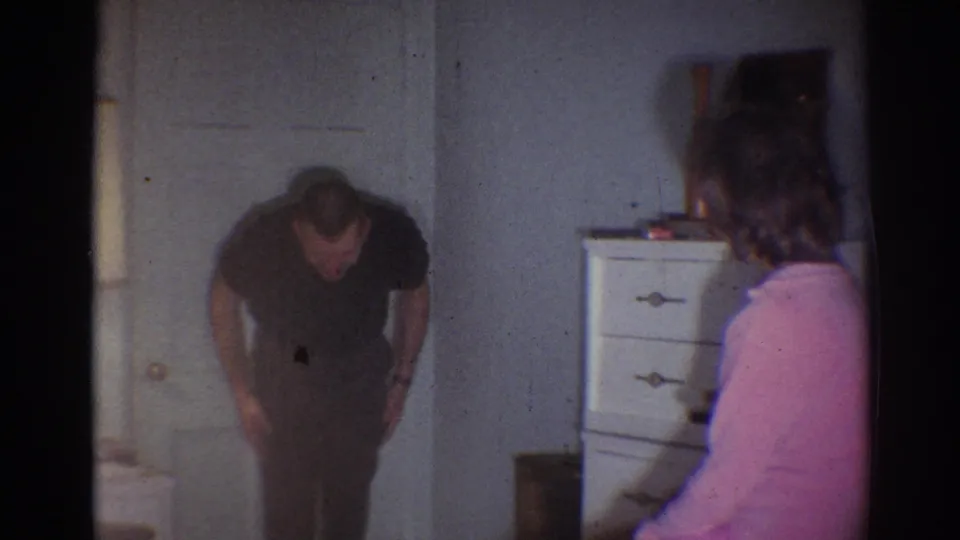 In a 1967 Los Angeles home-movie frame, a man bends forward in a dim room as a seated woman in pink watches from the side.